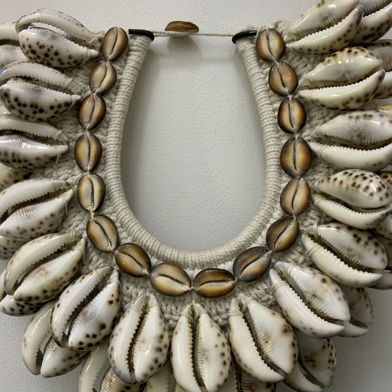 Handcrafted tribal-style necklace featuring polished Tiger Cowrie shells and money cowries woven onto a white macramé base, displayed on a black metal stand