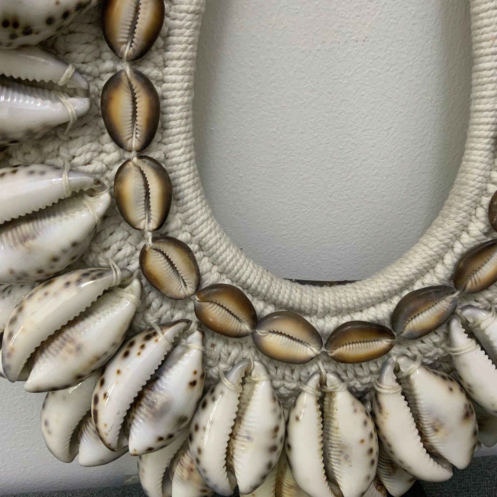Handcrafted tribal-style necklace featuring polished Tiger Cowrie shells and money cowries woven onto a white macramé base, displayed on a black metal stand