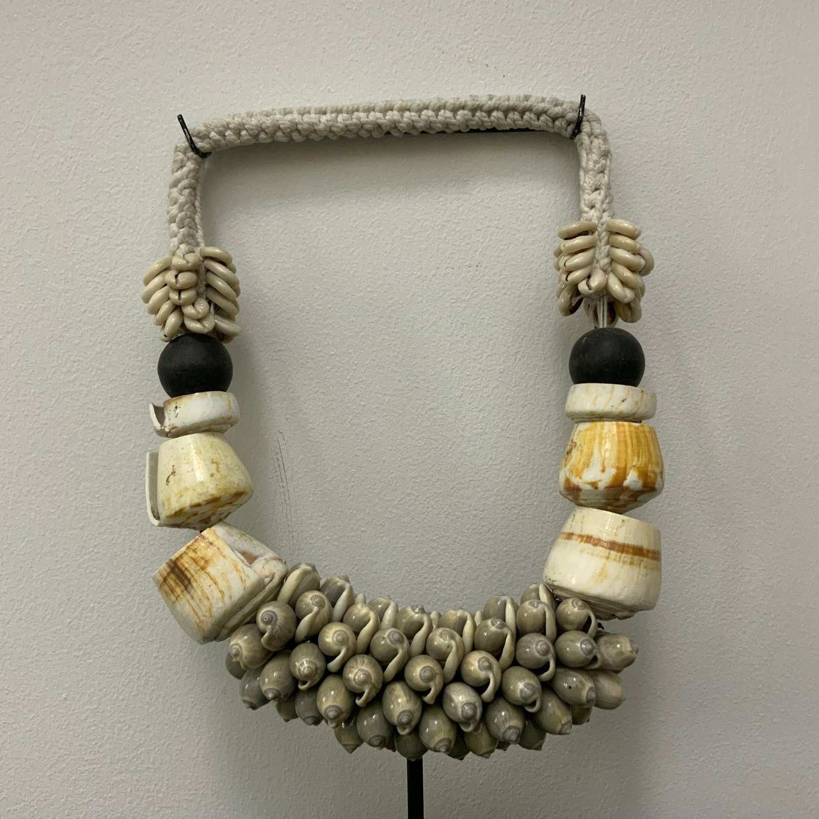 A handcrafted tribal-style statement necklace made of white sea shells, polished bone-colored resin rings, and black beads, displayed on a minimalist black metal stand